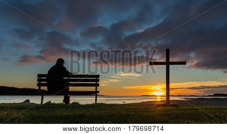 A man on a seat next to a cross watching the sunset at Foxton Beach river.