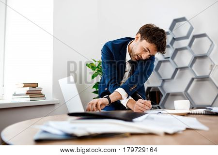 Absentminded young businessman in suit speaking on phone at workplace, writing, printing, office background.