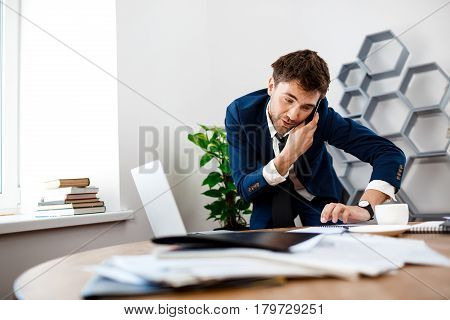 Absentminded young businessman in suit speaking on phone at workplace, smiling, office background.
