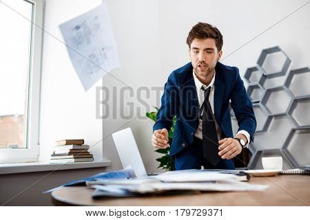 Absentminded young businessman in suit rummaging in papers at workplace, office background.