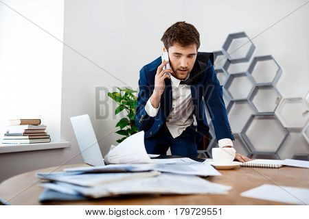 Absentminded young businessman in suit speaking on phone at workplace, office background.