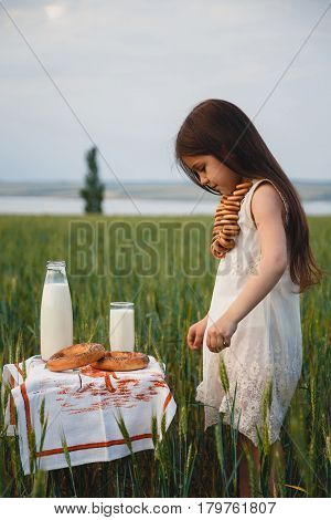 Cute little girl in white dress standing near small table with milk and bread on it in green field. Summertime.