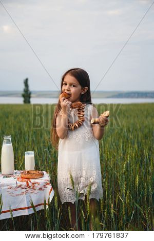Cute smiling little girl in white dress eating bagel with milk in green field summertime