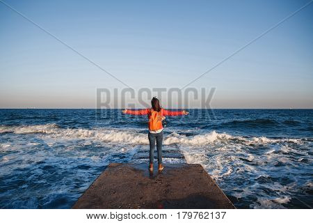 Young happy girl with orange backpack standing on a seaside breathing fresh air raising arms enjoying the view