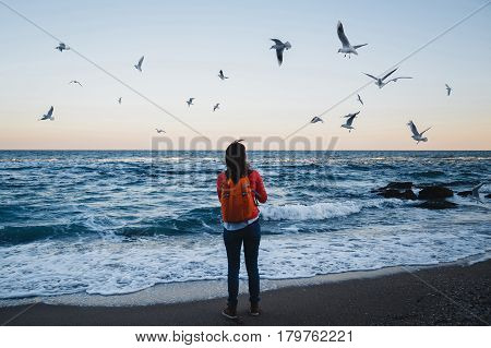 Young happy girl with orange backpack feeding seagulls on a seaside evening time