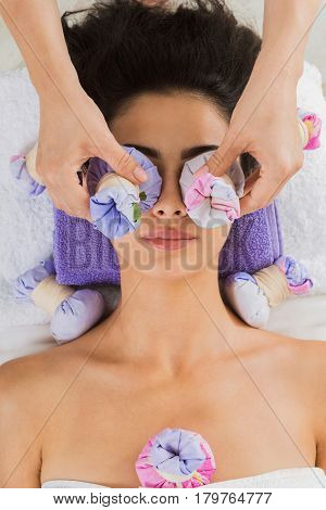 Herbal ball massage in ayurvedic spa. Female massagist with young woman in wellness center. Healthcare therapy to beautiful indian girl in beauty parlor, top view of face with eyes closed