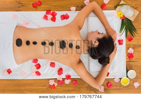 Black marble stone massage in spa. Top view of beautiful girl with stones on back and rose petals in wellness center. Pretty woman relaxation procedure, attractive young indian girl in beauty parlor.