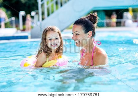 Young beautiful mother in bikini playing with her daughter in inflatable ring in swimming pool in aqua park. Summer heat and water.