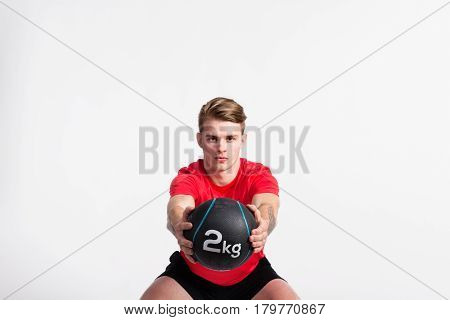 Handsome hipster fitness man in red t-shirt holding medicene ball, doing squat. Studio shot on gray background.