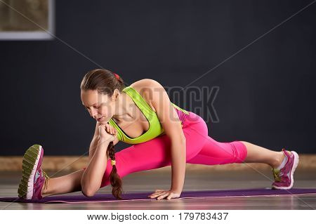 Woman practicing yoga in the splits position on the floor. In the gym. Sporty woman in the sporty clothes, pink leggings and green t-shirt. Healthy lifestyle. Portrait of the yoga woman on the mat.