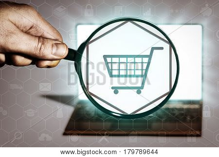 Hand holds the magnifying glass in front of an open notebook. Among the many icons attention is focused on the cart icon. Search for goods online shopping stores buy.