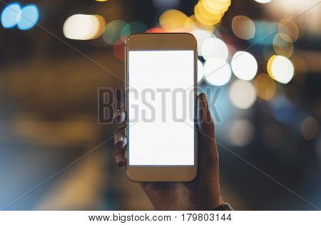 Girl pointing finger on blank screen smartphone on background illumination glow bokeh light in night atmospheric christmas city hands using template mobile phone on lights auto and glitter street; mockup