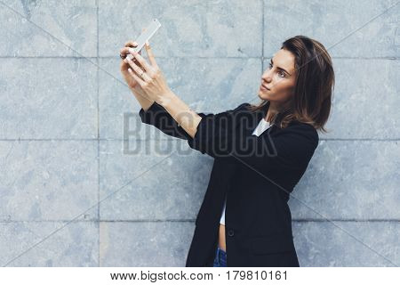 Portrait young businesswomen in black suit using smartphone isolated on background concrete gray wall mockup pretty hipster manager holding mobile gadget girl making selfie photo connect