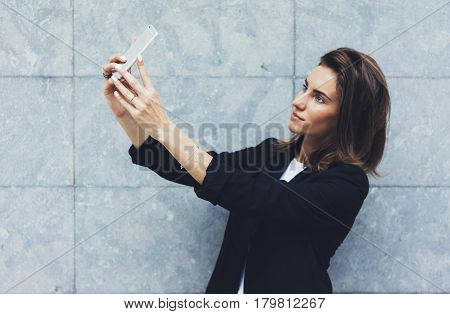 Portrait young businesswomen in black suit using smartphone isolated on background concrete gray wall mockup pretty hipster manager holding mobile gadget girl making selfie photo connect