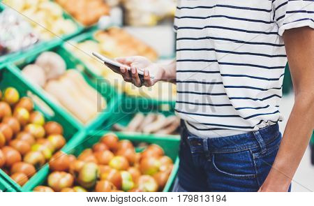Young woman shopping healthy food in supermarket blur background. Female hands buy products tomato using smartphone in store. Hipster at grocery using smartphone. Person comparing price of produce