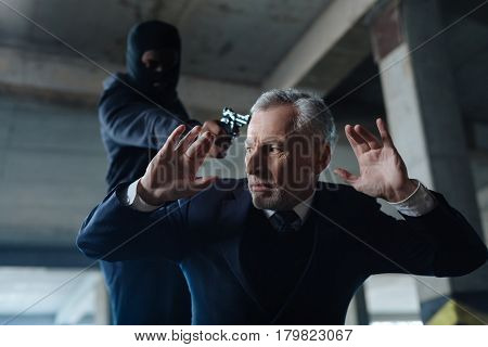 Abduction victim. Unhappy frightened aged man standing on his knees and raising his hands while being abducted