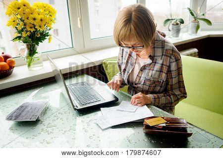 The elderly woman works behind the laptop. On a table various receipts and accounts lie. Modern pensioners use modern technologies.