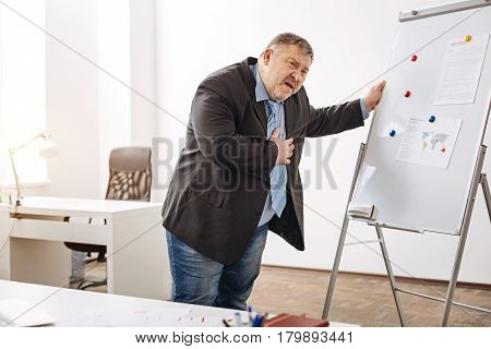 What is that feeling. Tired helpless overloaded man standing by the pin board and holding his chest while feeling pain there