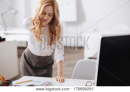 Straggling with horrible pain . Disappointed tired young office manager standing in the office and working while touching her back and suffering from ache