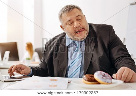 Watering mouth. Immoderate compulsive witty guy feeling strong desire of taking a bite of a doughnut lying on his desk