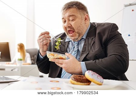 Imagining tastier things. Stout unhealthy office worker eating a salad trying being healthy but still wanting eating some junk food
