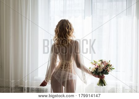 Bride near the window in her underwear. Blond sexy female Boudoir photo back to camera in front of large window. Bridal bouquet in the hand of bride.