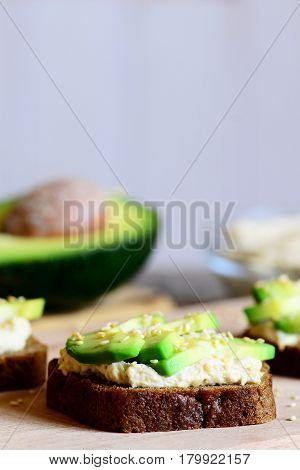 Hummus avocado sandwiches on a wooden board, avocado half, hummus in a glass bowl. Open sandwiches made with rye bread, avocado slices, hummus and roasted sesame seeds. Vertical photo
