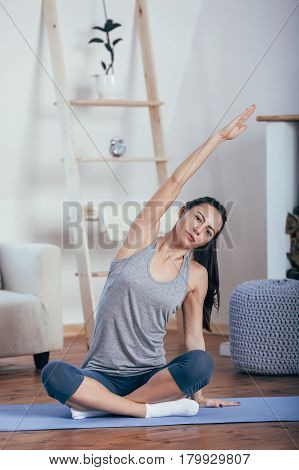Young cheerful attractive woman practicing yoga, sitting in Sukhasana pose, working out wearing sportswear, top, pants, indoor full length