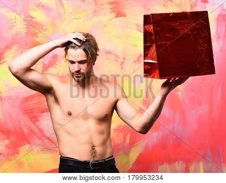 Bearded man short beard. Caucasian sexy macho man with moustache shows six packs and abs on muscle torso holding red package or bag on colorful studio background