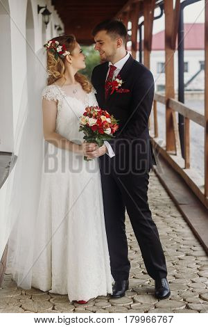 Happy bride hugging with groom beautiful blone bride in white wedding dress walking with handsome groom on paving road near wall happy newlyweds