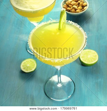 A square photo of a lemon Margarita cocktails with wedges of lime and a salted nuts snack, on a vibrant turquoise background with copy space