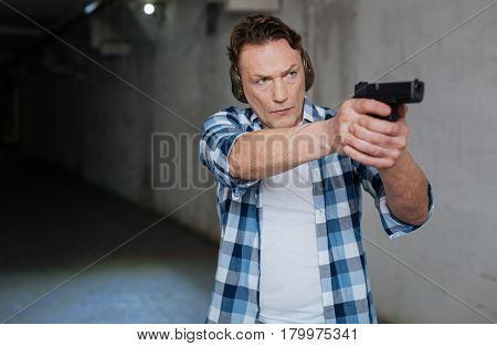 Professional marksman. Handsome nice professional marksman wearing headphones and holding a handgun while aiming at the target