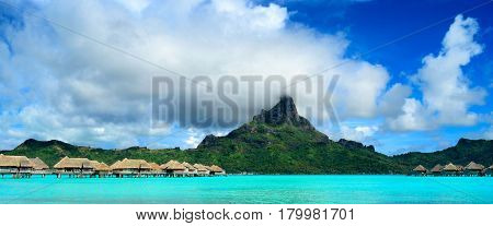 Panorama image of a tropical Bora Bora landscape with green Otemanu mountain behind a luxury resort in the turquoise lagoon of the island near Tahiti in Pacific French Polynesia.