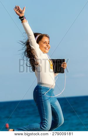 Close Up Action Portrait Of Smiling Tween Girl Jumping With Headphones And Tablet On Beach. Girl Rai