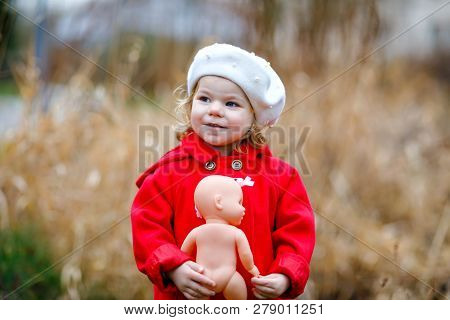 Outdoor Portrait Of Little Cute Toddler Girl In Red Coat And White Fashion Hat Barret With Toy Doll.