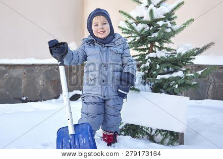 Cute Little Caucasian Baby Boy Shovels Snow In The Yard With Fir-tree On Background. Winter Outdoors