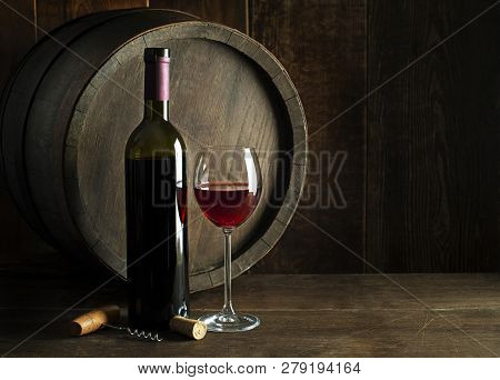 Bottle Of Red Wine With Glass On Wooden And Barrel Background. Glass Of Wine And Cork.