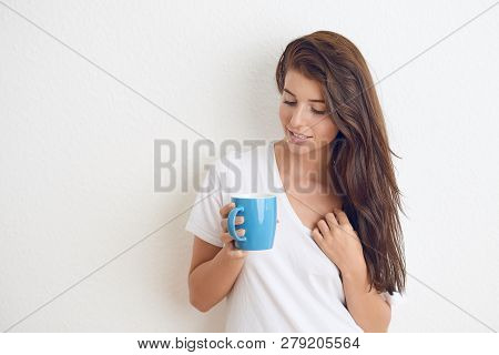 Young Brunette Woman In White T-shirt Drinking From Blue Mug With Both Hands, Looking At Camera With