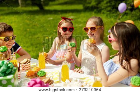 holidays, childhood and celebration concept - happy kids in sunglasses sitting at table on birthday party at summer garden and eating cupcakes