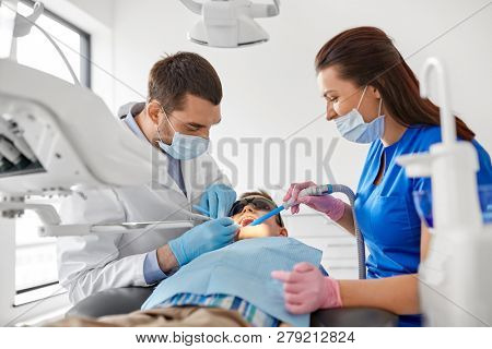 medicine, dentistry and healthcare concept - dentist and assistant with dental drill and saliva ejector treating kid patient teeth at dental clinic
