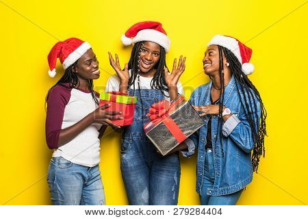 Three African Woman In Santa Hat Share Christmas Present On Yellow Background