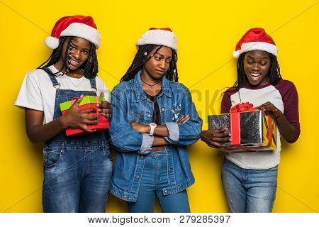 Three African Woman In Santa Hat Share Christmas Present On Yellow Background