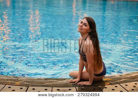Sexy Woman Sitting On The Edge Of Swimming Pool, Wearing Bikini While On Vacations In A Sunny Tropic