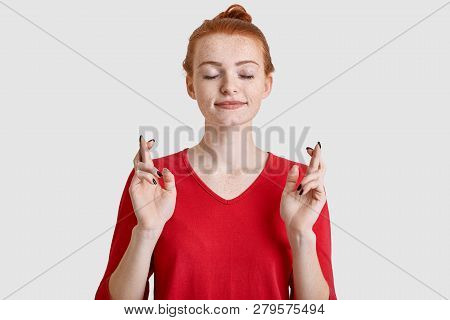 Photo Of Freckled Young Woman With Minimal Make Up, Has Ginger Hair Combed In Knot, Crosses Fingers 