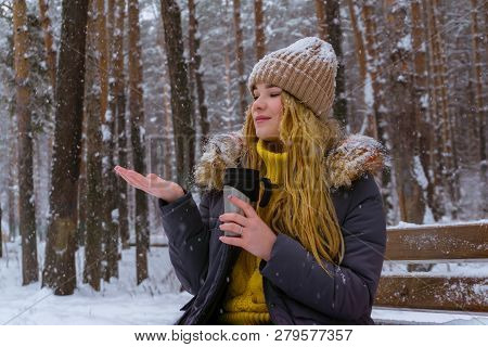 Girl Holding A Thermomug And Catches Snowflakes With Her Hand While Sitting On A Bench In The Winter