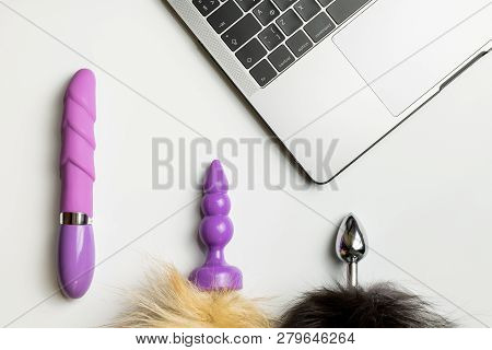 Dildo, Anal Plug Tails And Laptop On White Desk. Online Dating For Casual Sex, Promiscuity And Polyg