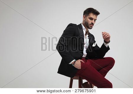 portrait of seductive fashion man sitting on wooden chair on light grey background with a hand in pocket