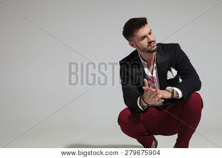 portrait of fashion man squatting on light grey background  and looking to side while holding hands together