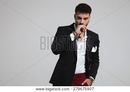 portrait of relaxed fashion man coughing while standing on light grey background with hand in pocket and holding hand to mouth
