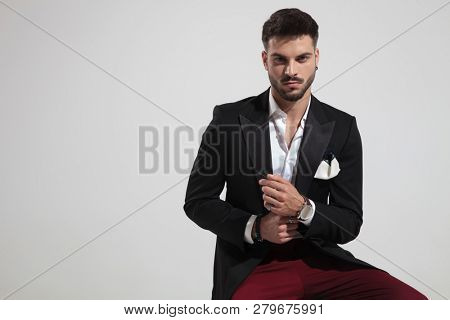 portrait of seated elegant man wearing red pants and black suit fixing his watch on light grey background